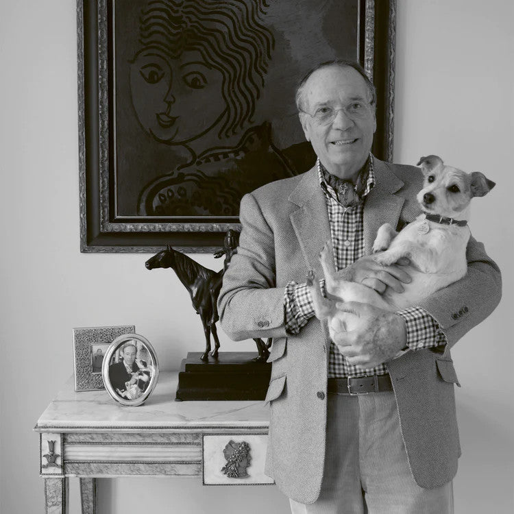 a smiling olivier creed holding his dog whilst standing in front of a painting and a desk
